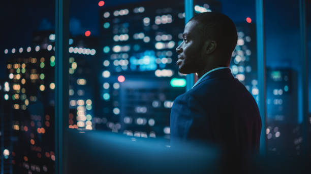 Successful Businessman Looking Out of the Window on Late Evening. Modern Hedge Fund Investor Enjoying Successful Life. Urban View with Down Town Street with Skyscrapers at Night with Neon Lights. exitoso hombre de negocios mirando por la ventana a última hora de la noche. inversor de fondos de cobertura moderno que disfruta de una vida exitosa. vista urbana con calle del centro de la ciudad con rascacielos por la noche con luces de neón.