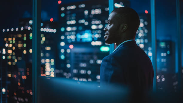 successful businessman looking out of the window on late evening. modern hedge fund investor enjoying successful life. urban view with down town street with skyscrapers at night with neon lights. - stad fotos stockfoto's en -beelden