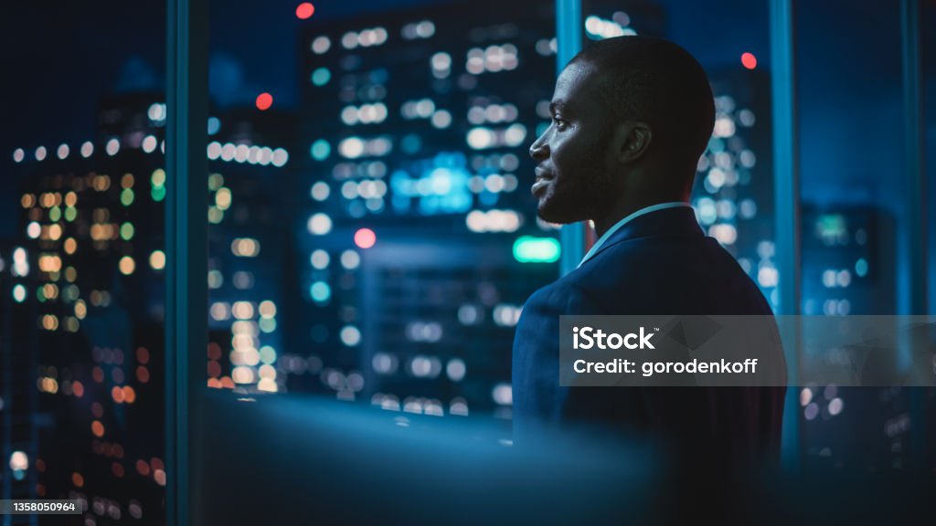 Successful Businessman Looking Out of the Window on Late Evening. Modern Hedge Fund Investor Enjoying Successful Life. Urban View with Down Town Street with Skyscrapers at Night with Neon Lights. Business Stock Photo