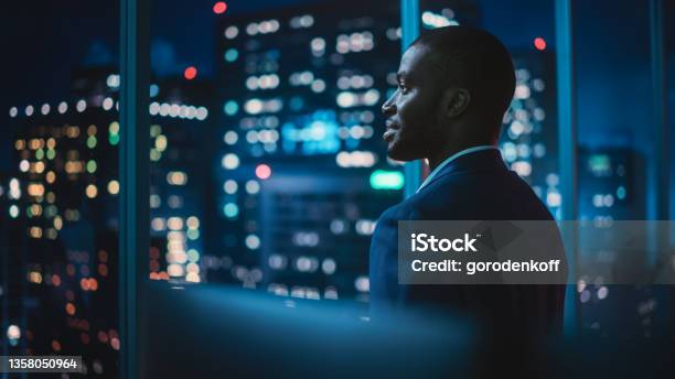 Successful Businessman Looking Out Of The Window On Late Evening Modern Hedge Fund Investor Enjoying Successful Life Urban View With Down Town Street With Skyscrapers At Night With Neon Lights Stock Photo - Download Image Now