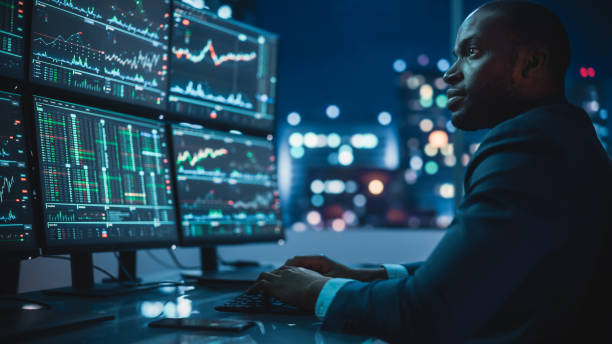 istock Financial Analyst Working on Computer with Multi-Monitor Workstation with Real-Time Stocks, Commodities and Exchange Market Charts. African American Businessman Works in Investment Bank Late at Night. 1358050967