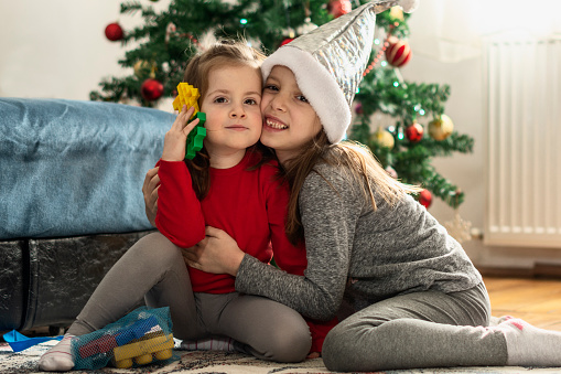 Two girls embracing while sitting in front of Christmas tree