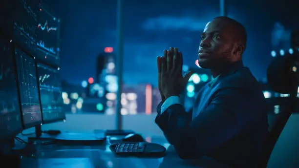 Photo of Financial Analyst Working on Computer with Multi-Monitor Workstation with Real-Time Stocks, Commodities and Exchange Market Charts. African American Trader Works in Investment Bank Late at Night.