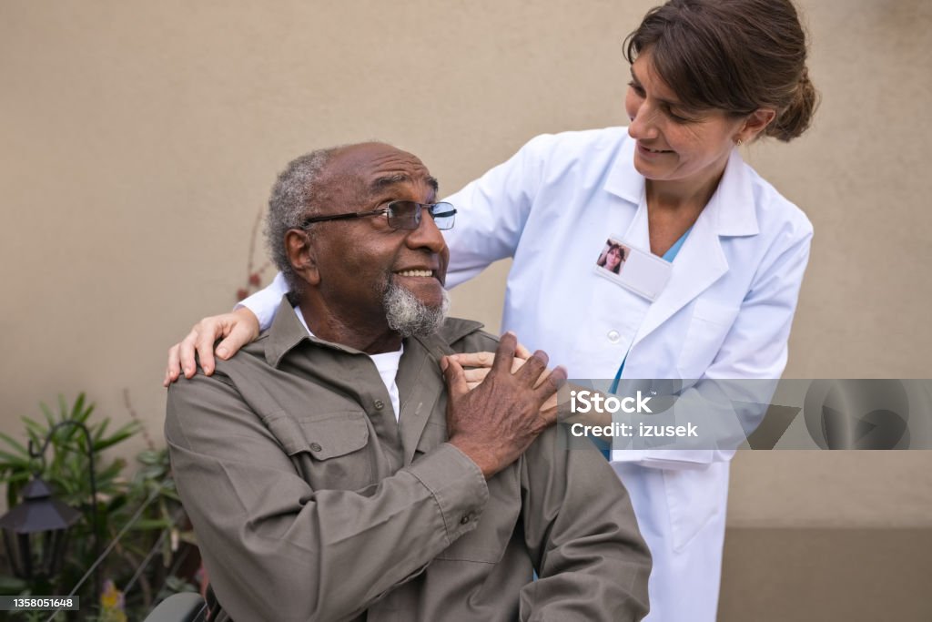 Caregiver talking with senior man against wall Happy female nurse talking with senior man. Elderly male is looking at caregiver. She is taking care of patient against wall. Hand On Shoulder Stock Photo