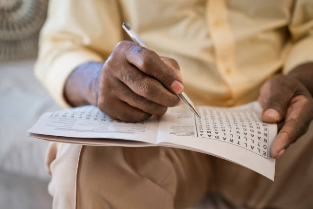 Elderly man solving puzzle at home Senior man solving word search puzzle at home. Elderly male is with pen and book. He is sitting in living room. crossword stock pictures, royalty-free photos & images