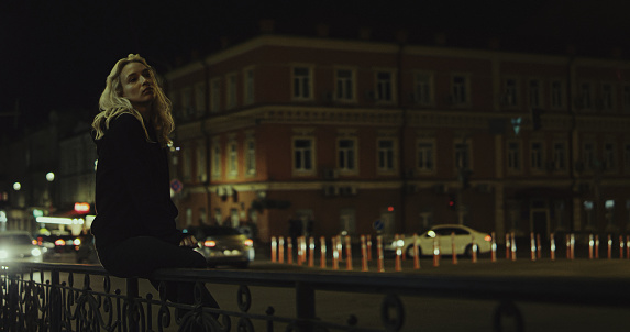 Beautiful woman sitting on bench while cars passing in city lights street. Thinking female person with blonde hair looking city view in urban area. Thoughtful girl spending time at town.