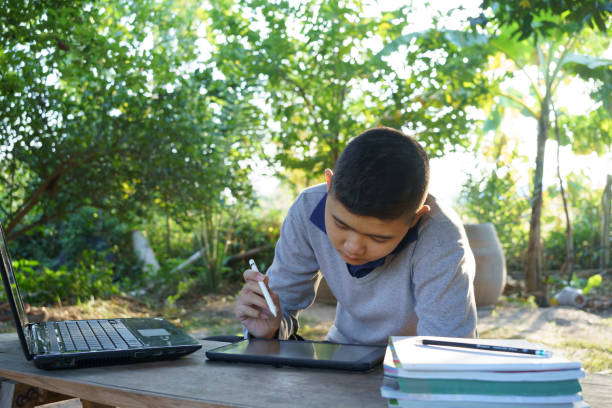istock Boy is studying online on a wooden table with a laptop computer and tablet during morning hours in a rural home. concept online education countryside area and work from home 1358052639