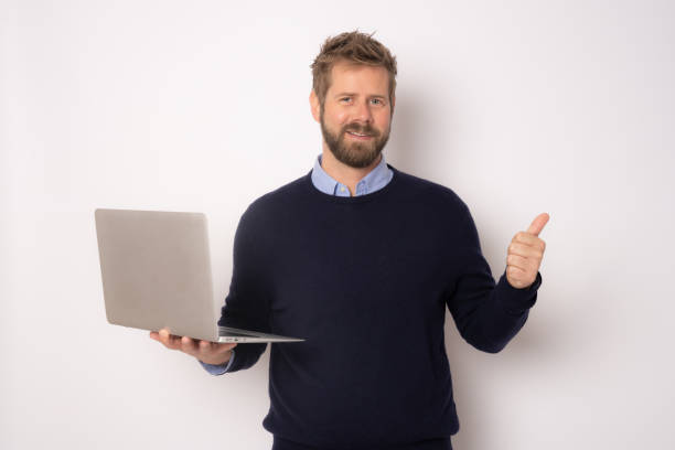 istock Young smiling handsome man holding laptop computer with thumb up standing isolated over white background. 1358054335
