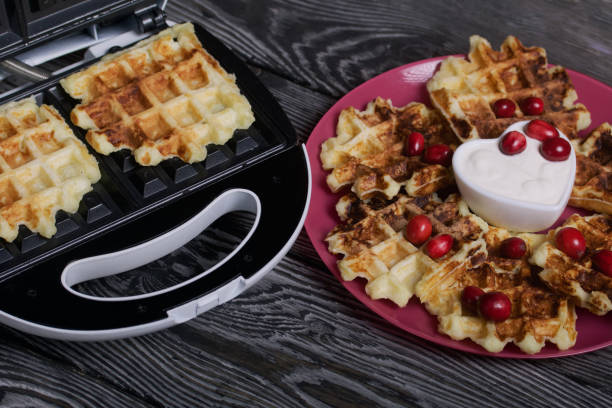 istock Waffles with sour cream and cranberries. Lie on a plate. Next to it is a waffle maker with waffles. On black pine boards. 1358057093