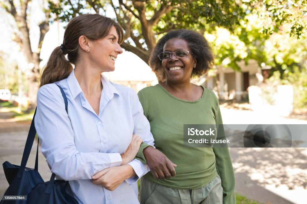 Senior woman and caregiver talking on footpath Smiling senior woman talking with healthcare worker. Nurse and elderly female is with arm in arm. They are standing on footpath. Women Stock Photo