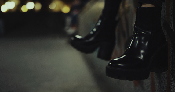 Female person moving legs in black shoes while sitting on urban building in night city lights. Unknown woman making movements in high heel shoes. Close up woman legs in city walk night.