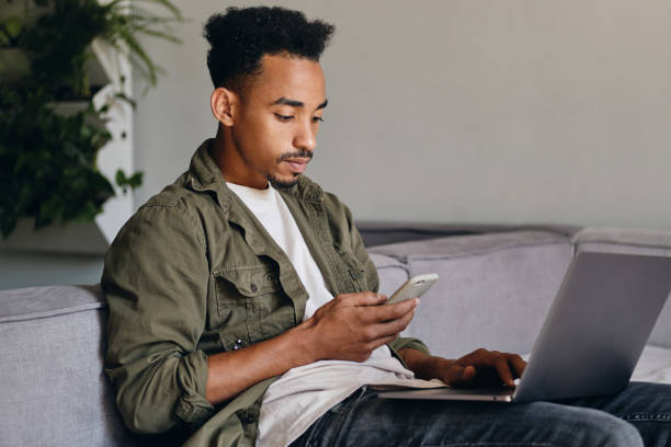 istock Young serious African American guy with laptop thoughtfully using cellphone in modern co-working space 1358062312