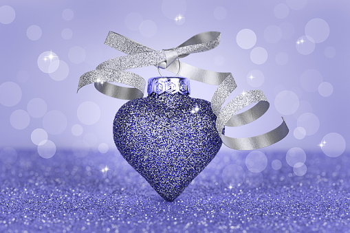 Christmas toy in the form of a heart on a violet bokeh background. Christmas card. Sparkling texture. Very peri, color of the year 2022.