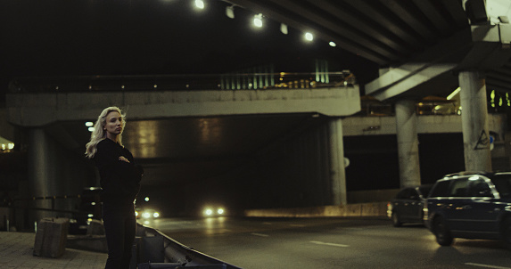 Young woman standing on dark road while cars driving under bridge in night city lights. Beautiful female person with blonde hair looking camera on highway. City girl enjoying life at night.