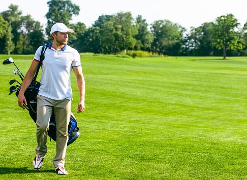 young male golfer carries a bag of golf clubs. Slim man in white T-shirt , golf shoes, white cap and beige pants is going in the green Turf Grass. Copy Space. Full-length. High quality photo