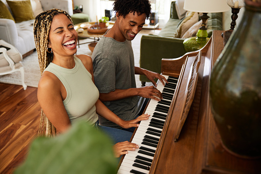 Laughing young woman playing a duet with her boyfriend on a piano in their living room at home