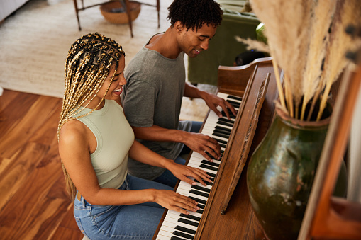 Smiling young couple sitting on a bench and playing a duet together on a piano in their living room at home