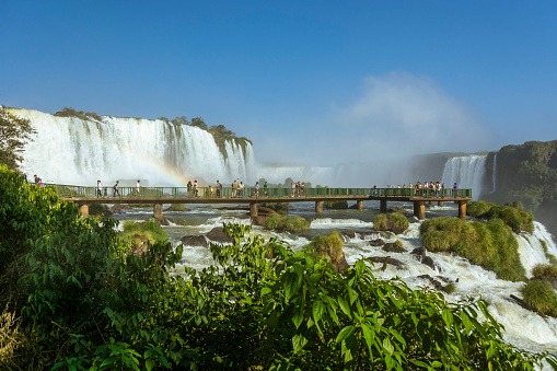 Foz do Iguaçu, Brazil: Tourists visiting the Iguaçu Falls through the observation bridges on the Brazilian side. Panoramic view of waterfalls on a sunny day and blue sky.