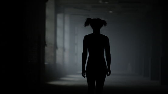Silhouette of female athlete walking in corridor. Back view sporty woman going in dark loft building. Afro fit girl resting after cardio training. Muscular trainer wearing sportswear