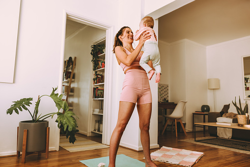 Cheerful mom smiling at her baby while lifting her up. Happy young mom working out with her little baby at home. New mom bonding with her baby during her post-natal fitness routine.