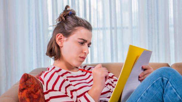 chica sentada en el sofá, estudiando con cuaderno amarillo en la mano, una joven con medio pedazo de pelo. tiene una sudadera a rayas rojas. escribe en su cuaderno con el bolígrafo que sostiene. - raised eyebrows women human face thinking fotografías e imágenes de stock