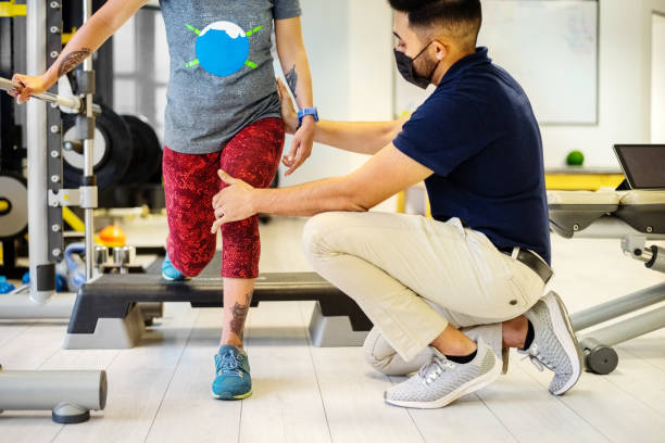 Physiotherapist with face mask helping woman with cerebral palsy do lunges at rehab Cropped shot of a physiotherapist wearing face mask helping woman in doing legs stretches at rehab gym during pandemic checking sports stock pictures, royalty-free photos & images