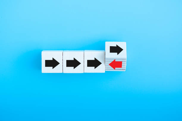 istock Red arrow flip over wooden cube block with red arrow opposite directon black arrows think different and standing out from the crowd concept isolated on blue background. 1358089083