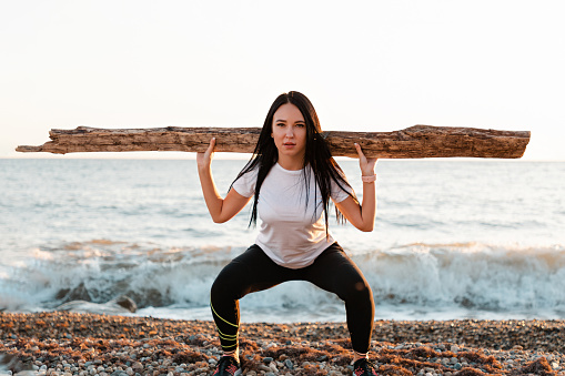 The concept of sport. Young brunette squats with a log on her shoulders. In the background, the sea and the horizon line.