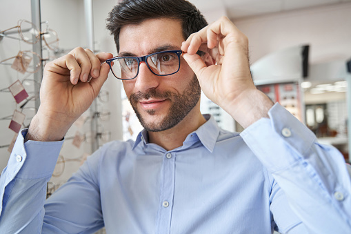 Pleased man in optician shop holding eyeglasses on his face by sides while staring forward