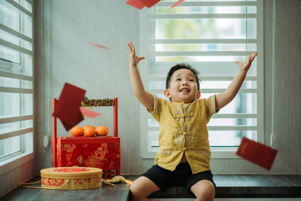 Asian chinese smiling cutie boy sitting and throwing up red envelope in living hall during chinese new year stock photo
