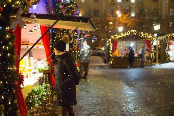 A woman looks and buys at a kiosk, people walk at the Christmas market in the festive decor of the streets. Exhibition and shopping malls in the city center. New Year. stock photo