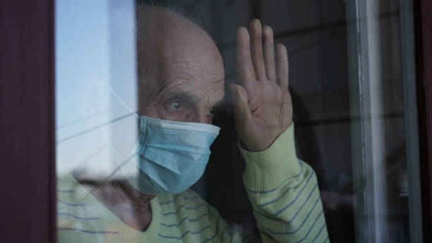 istock Lonely mature grandfather in facemask looking through closed window 1358101298