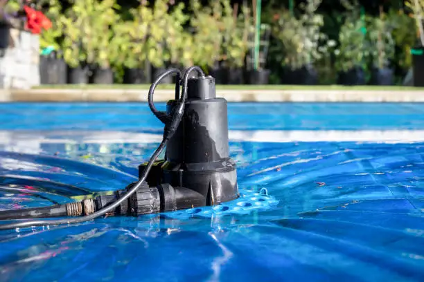 Photo of Automatic pool cover pump on top of blue wet cover in front of defocused garden foliage.