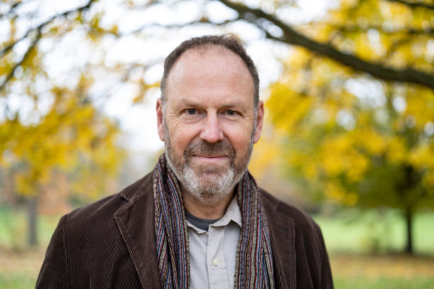 Headshot of 55 year old Caucasian man amidst fall foliage Front view of man with brown hair and gray beard standing in natural parkland wearing warm clothing and smiling at camera. 50 59 years stock pictures, royalty-free photos & images