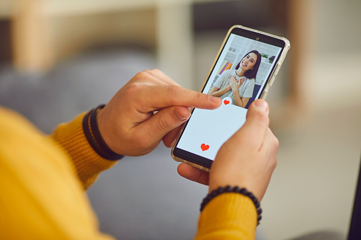 Dating app or site in mobile phone screen. Close up of a lonely man uses a smartphone to flip through women's profiles and presses a heart button with his finger. Banner or mockup website concept.
