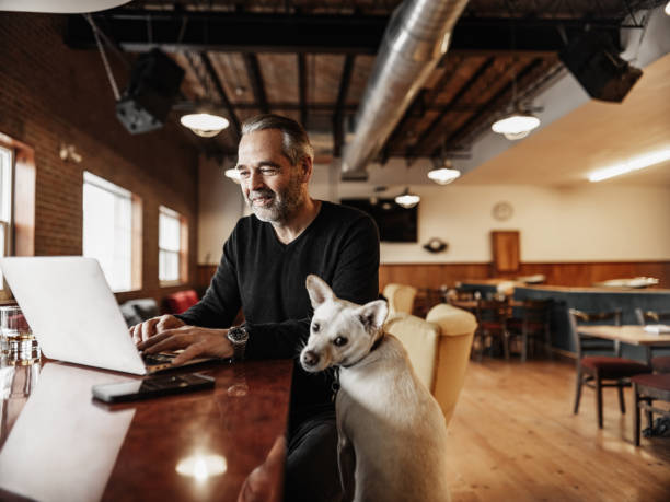 Owner of small business with his small dog working at the bar Owner of small business with his small dog working at the bar ,  working on lap top while having a drink. Dressed in casual clothes with black sweater and jeans. Focus on a small dog in the foreground.  Interior of vintage bowling alley in the morning with natural light. dog disruptagingcollection stock pictures, royalty-free photos & images