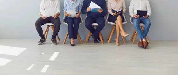 Recruitment process and office paperwork concepts. Low section crop of group of job candidates waiting in line for interview. Company workers with documents sitting in queue on chairs