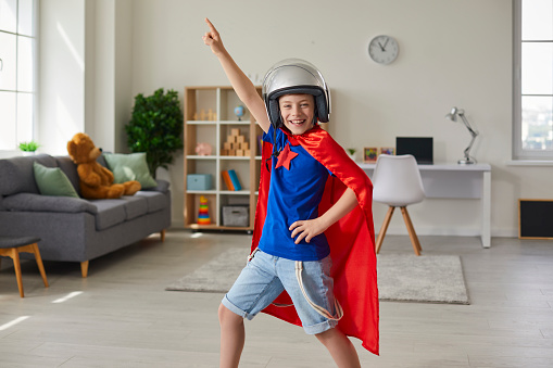 Confident cheerful 10 year old boy in superman costume having fun in nursery room looking at camera. Happy smiling kid in helmet and red superhero cape playing and dancing enjoying fun games at home