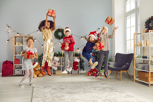 Multiracial kids enjoying winter holidays and wonderful New Year gifts. Group of happy excited diverse children having fun and jumping for joy with their Christmas presents at cozy party at home