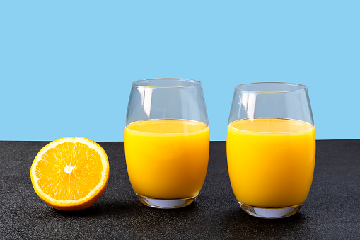 two glasses with orange juice, half of orange fruit next to glass as drink, on blue background