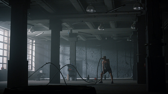 Wide shot powerful man battling ropes in gym. Athletic man doing fitness workout in loft building. Fit sportsman training with battle ropes. Male bodybuilder exercising with sports equipment