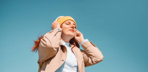 image d’une jolie jeune femme rousse caucasienne souriant en écoutant de la musique ou un podcast à l’aide d’écouteurs sans fil sur fond de ciel bleu. profiter du concept de voyage et de nature - headphones relaxation outdoors caucasian photos et images de collection