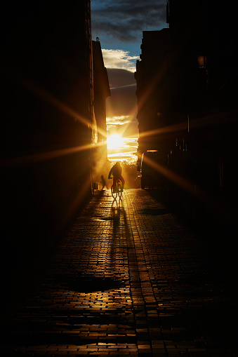 The silhouette of a cyclist outlined against the light advances down a cobbled street illuminated by the setting sun. Vertical photography. Copy space.
