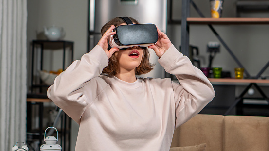 In the background, the kitchen and decorative items of the house are visible. Young girl in a light brown sweatshirt. She is holding vr glasses with his hands. Technology, simulation, hi-tech, videogame concept. She is playing games by feeling that she is alive while wearing virtual reality glasses. The video it's in is horrifying. She’s very surprised. Metaverse VR virtual reality game playing, Metaverse Concepts.