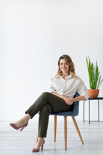 Happy middle-aged woman sits on chair. Minimalistic interior. White background with copy space.