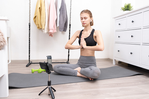 A slim girl is engaged in fitness online via a mobile phone. Young woman doing yoga and sports at home.