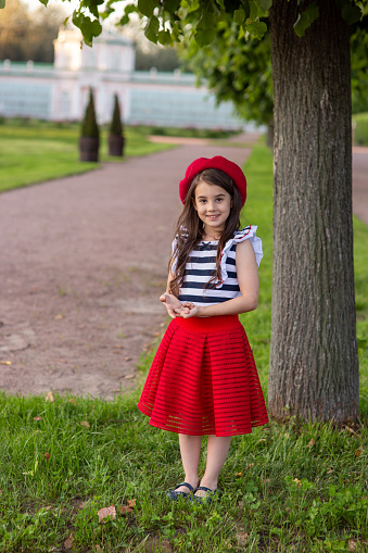 A beautiful little brunette girl, dressed in a French style: in a red beret, a striped T-shirt and a red skirt, stands under a tree in a summer park, smile, looks at the camera