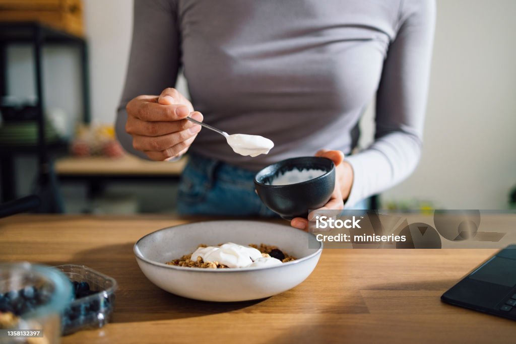 Close Up of Woman Hands Making Healthy Breakfast in Kitchen An anonymous Asian woman mixing cereals, greek yogurt and blueberries in a bowl on a kitchen table at home. Yogurt Stock Photo