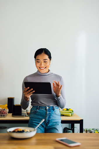 Happy Asian woman using digital tablet for online meeting while standing in a modern kitchen.