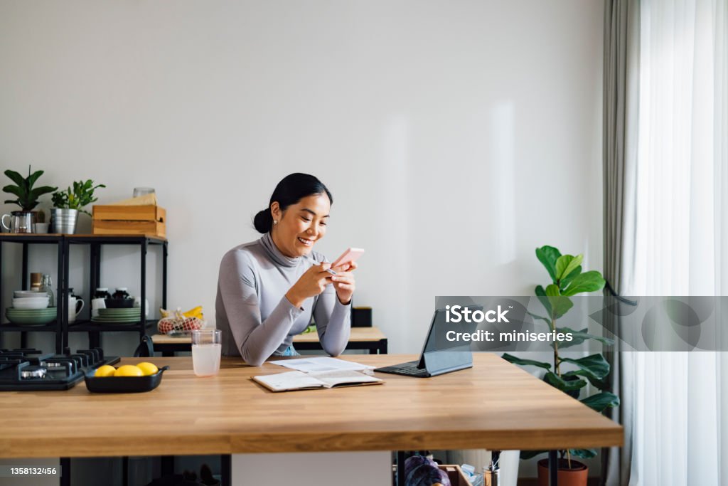 Happy Business Woman Working from Home Beautiful Asian businesswoman using digital tablet and notebook while typing text message on her mobile phone at kitchen desk. People Stock Photo